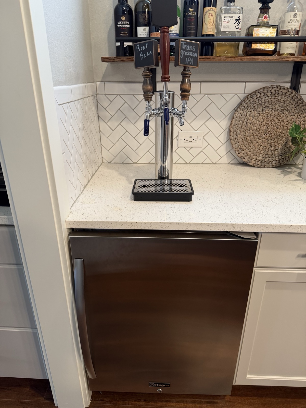 Kegco built-in kegerator under a bar counter, black stainless door with a keyed lock and curved handle, with a three-faucet chrome tap tower on the counter above and a wine/liquor bottle rack on the wall.
