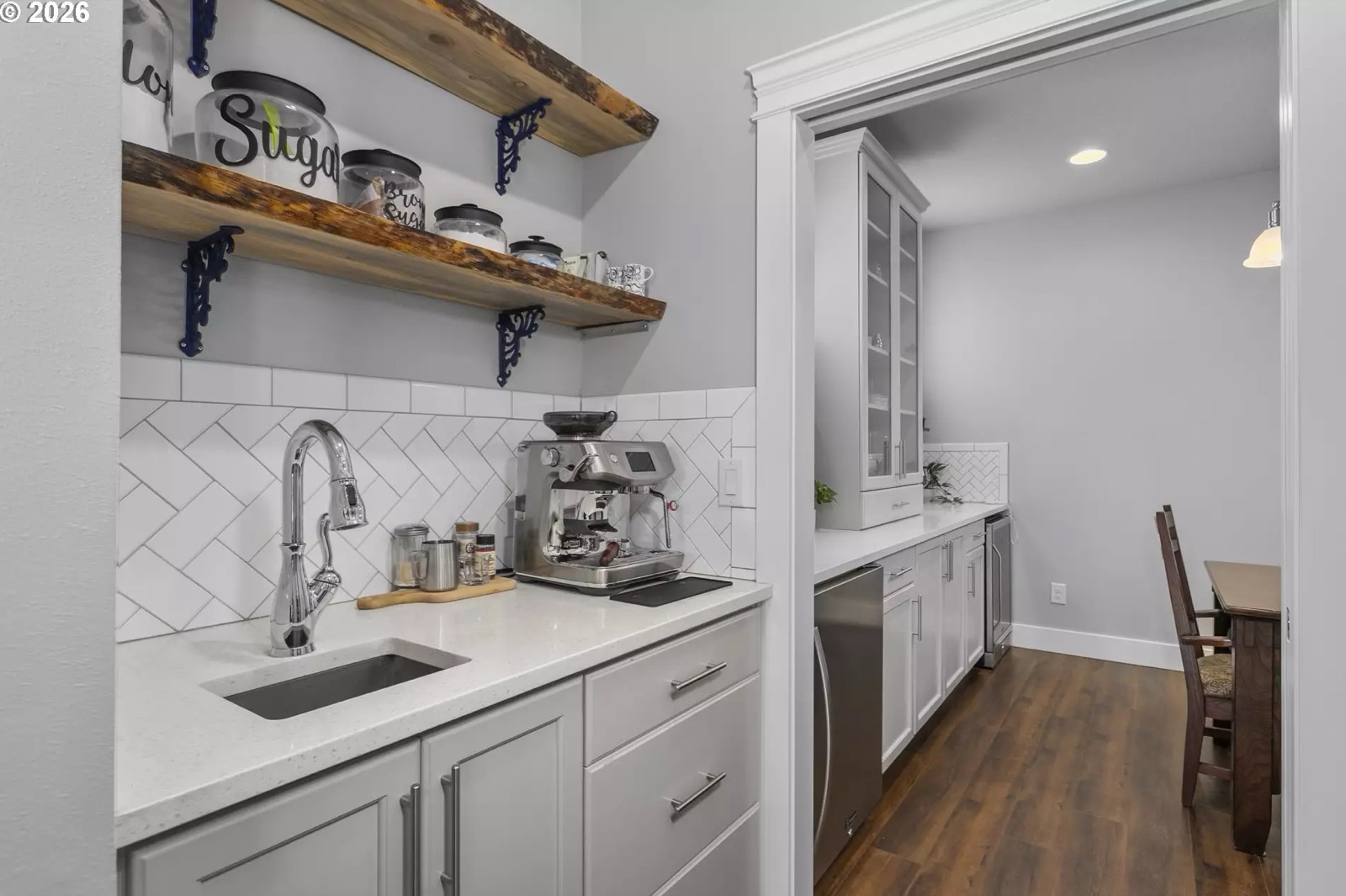 The butler’s pantry: a quartz counter with an undermount stainless sink and a pull-down chrome faucet, a chrome espresso machine beside it, two thick live-edge wood open shelves on cast-iron brackets holding glass canisters, a white herringbone tile backsplash, and a doorway at the right looking through to the dining room.