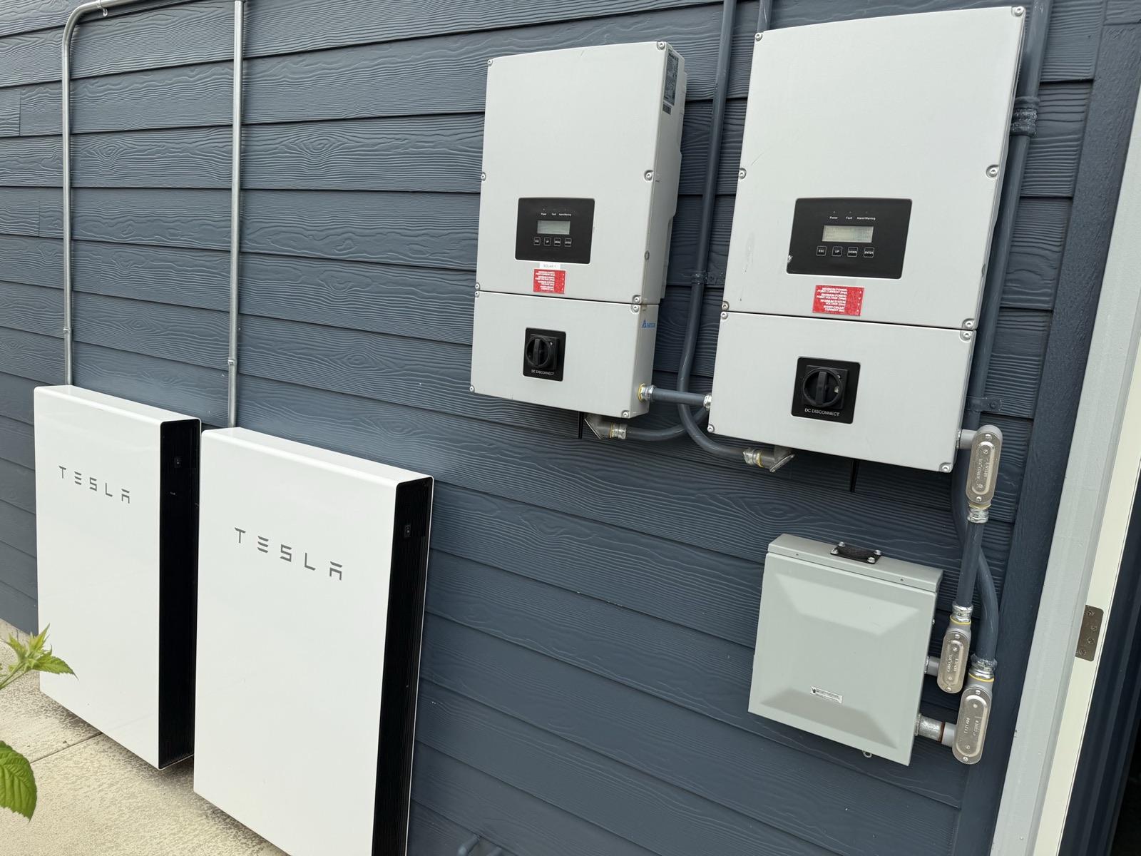 The two Tesla Powerwalls at ground level, the two Delta string inverters mounted on the wall above them, and a grey Square D-marked enclosure beside the inverters.