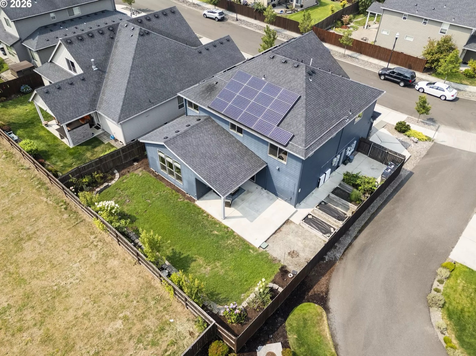 Aerial view of the whole property: the deep-navy two-storey house on its corner lot, the rooftop solar array, the covered patio and rear lawn, raised garden beds along the fence, the concrete driveway, and the street curving past.