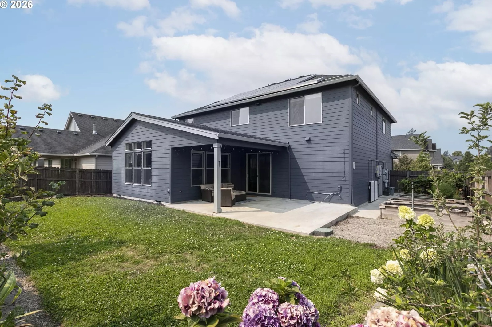 The rear of the house from the back lawn: deep-navy siding, a covered patio with seating and a sliding door, the solar array on the roof slope above, utility equipment mounted on the wall to the right of the patio, and raised garden beds and plantings at the right edge.