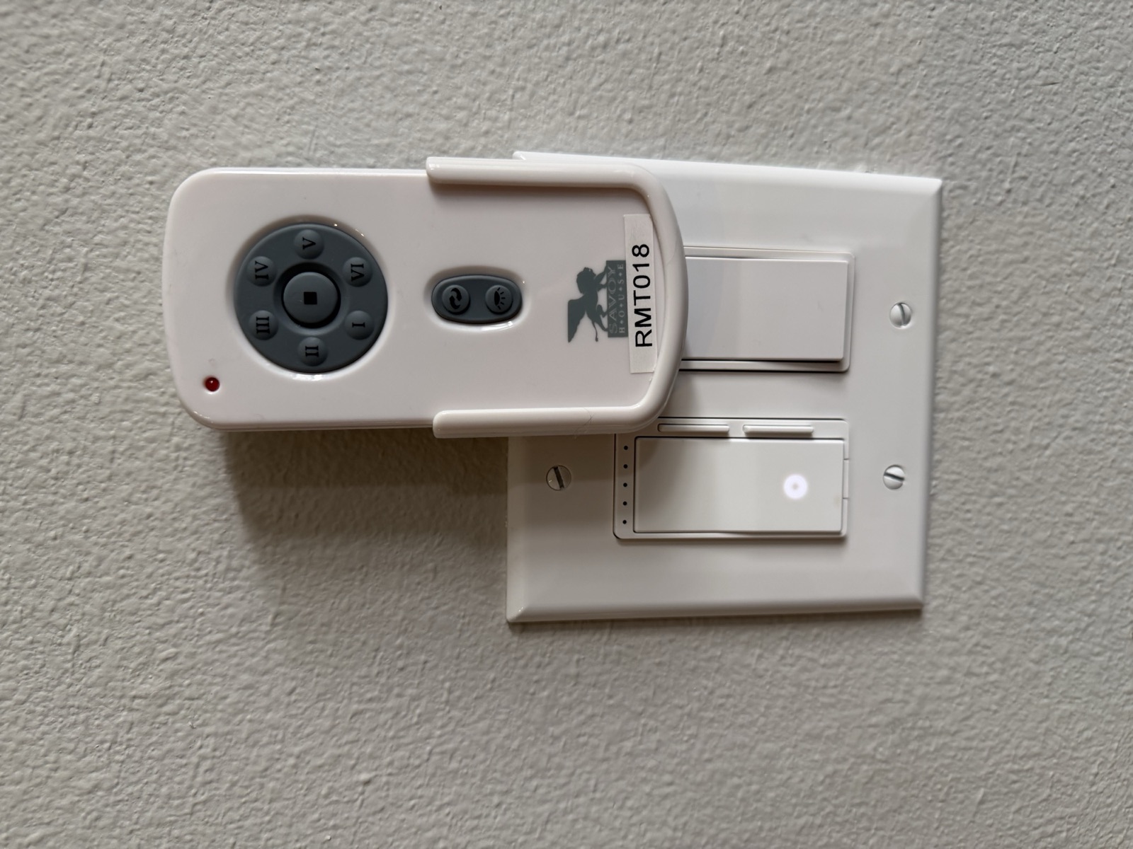 A Savoy House ceiling-fan handheld remote, labeled RMT018, sitting in its wall-mounted cradle next to a two-gang switch plate with an unlabeled paddle switch and a slide dimmer — the master bedroom fan's remote.