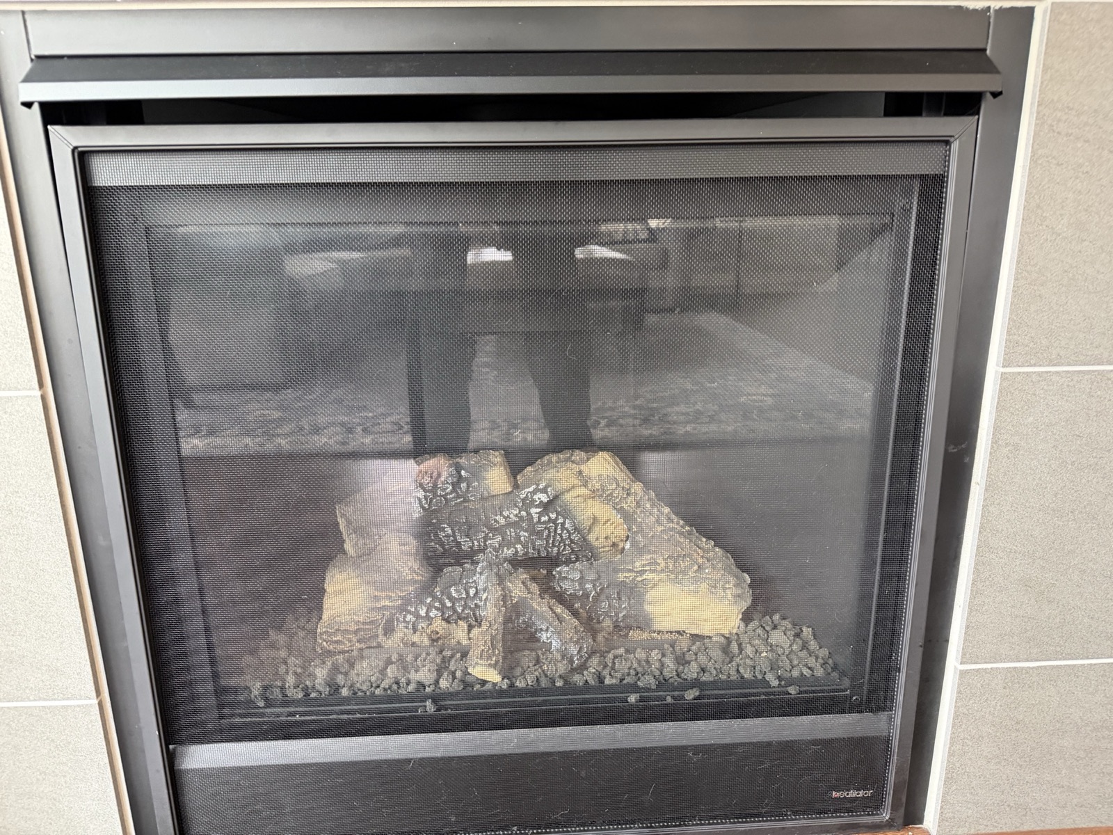 Direct-vent gas fireplace with ceramic logs and an ember bed behind a glass safety screen, set in a large-format grey tile surround; a Heatilator badge is visible at the lower right of the frame.