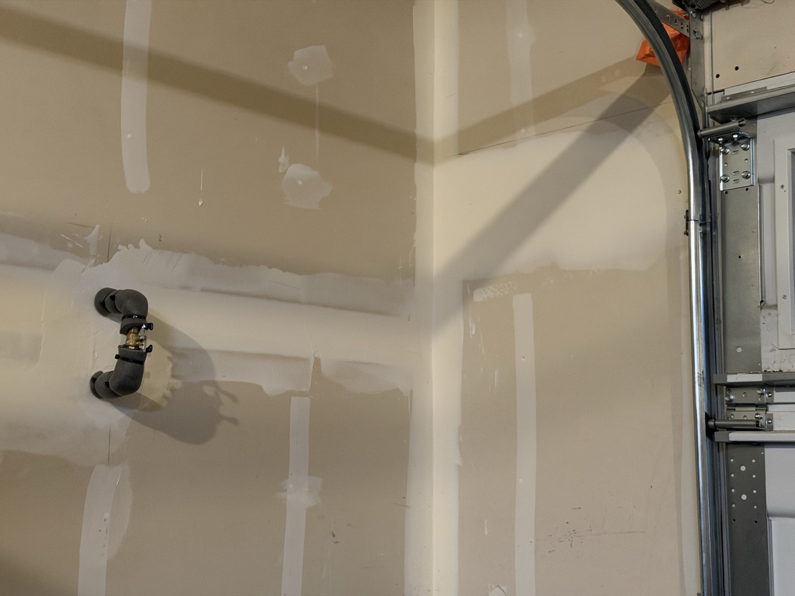A brass lever-handled ball valve set into black foam pipe insulation on a garage wall, immediately beside the overhead vehicle door track and hardware.