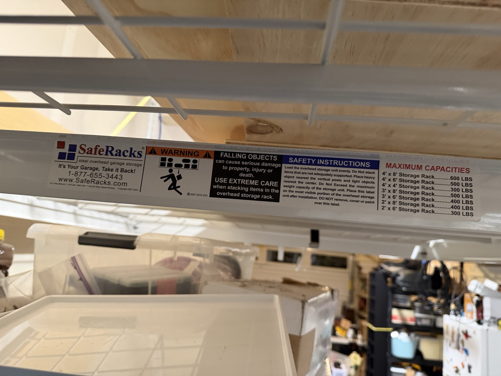 A white SafeRacks safety and capacity decal on a ceiling-mounted overhead storage rack rail, fully legible, listing maximum capacities from 300 to 600 pounds by rack size.
