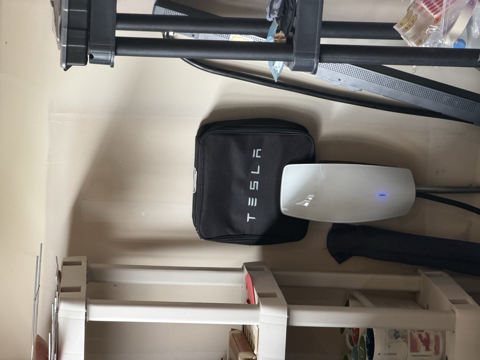 The white Tesla Wall Connector mounted on the garage wall, its single horizontal LED bar lit blue, with a black Tesla-branded Mobile Connector bag hanging on the wall beside it.