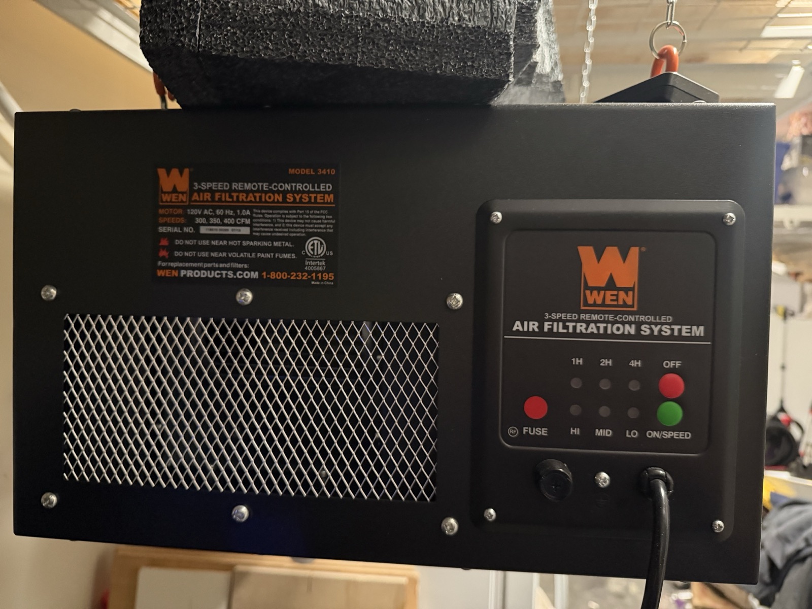 A black sheet-metal WEN 3410 shop air filtration unit hanging from ceiling chains, showing its nameplate, intake grille, and control panel.