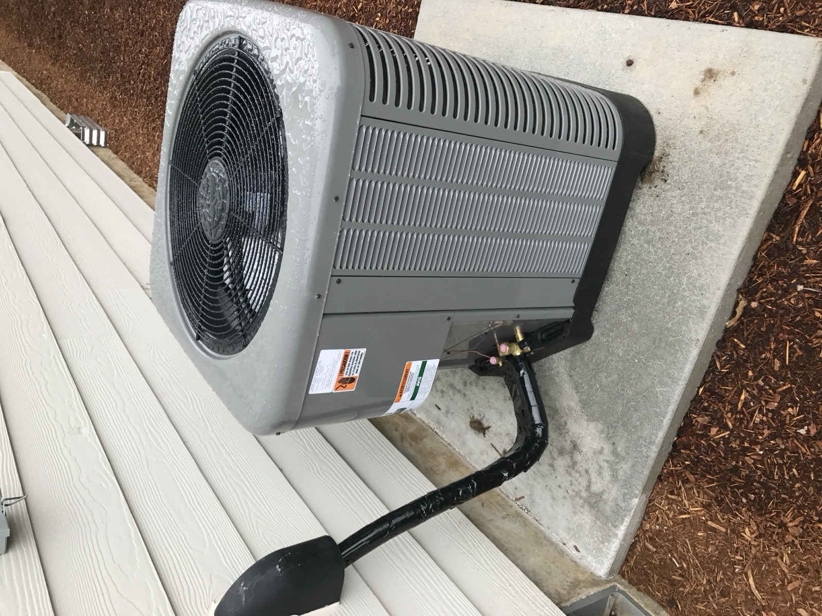 Outdoor Rheem condenser on its concrete pad in the side yard, freshly rained on, with the black-insulated line set entering the wall and the wall-mounted disconnect just above.