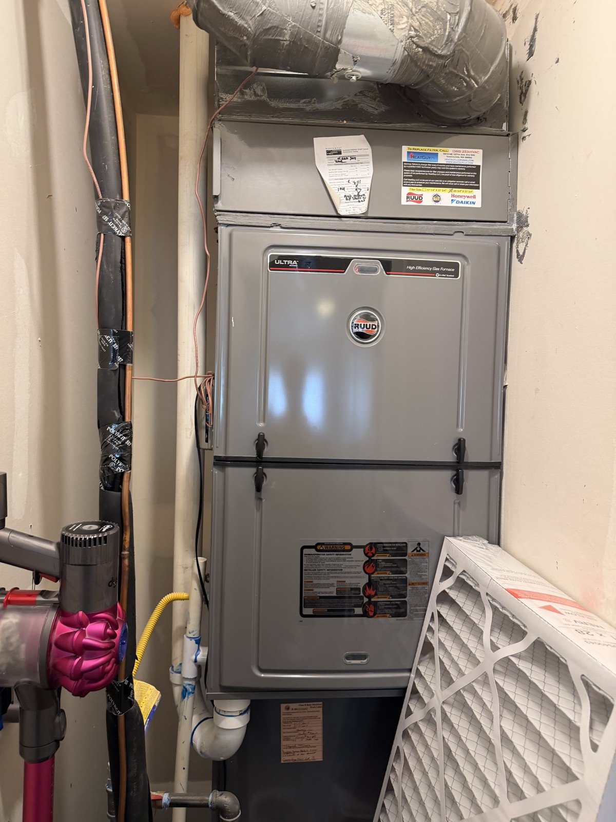 Ruud Ultra Series gas furnace in a tight utility closet, with a HeatGuy LLC service sticker on the filter cabinet, a spare MERV 8 filter leaning against it, and an insulated AC line set running along the floor.