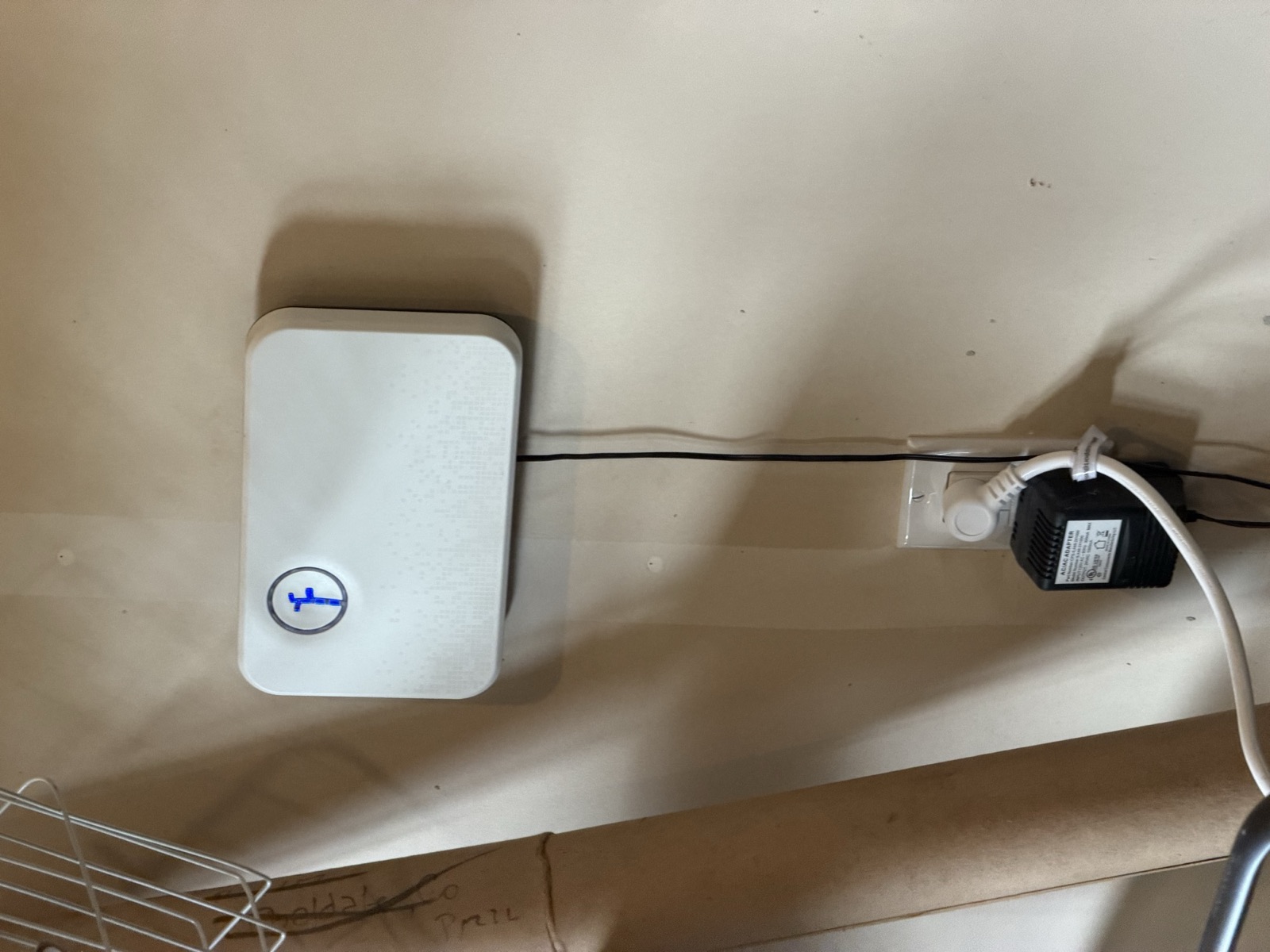 A white Rachio smart sprinkler controller mounted on an unfinished garage wall, with a single illuminated round button lit blue, powered by a black 24VAC plug-in transformer at a nearby duplex outlet.