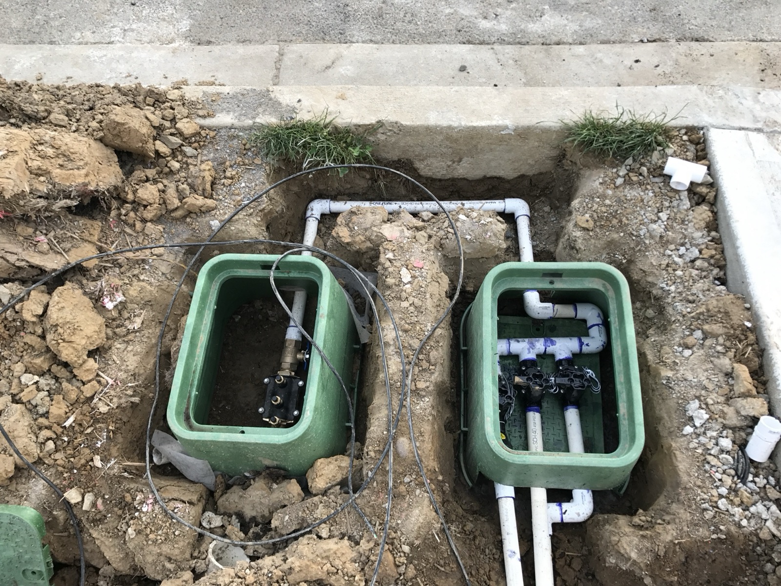 Two green irrigation valve boxes set in an open trench along the house’s poured concrete stem wall — the left box holding a brass-and-black-epoxy backflow assembly with ball-valve shutoffs, the right box holding three black solenoid valves on a common PVC manifold.