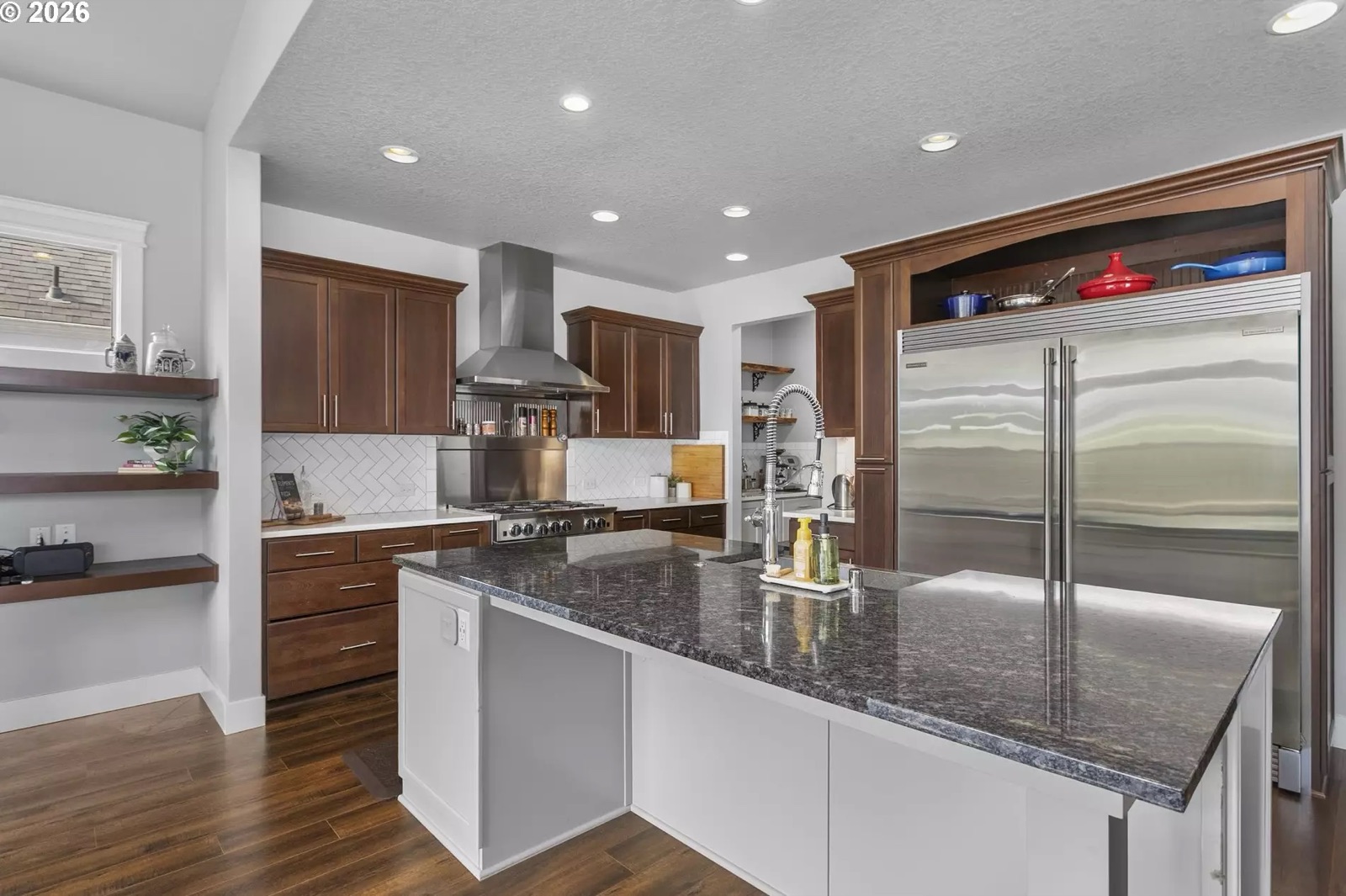 Wide view of the kitchen: a stainless BlueStar range under a stainless chimney hood against a herringbone tile backsplash, dark wood cabinetry, a long granite island with a prep sink and pull-down faucet, and a pair of full-height stainless refrigerator/freezer columns under a wood surround.