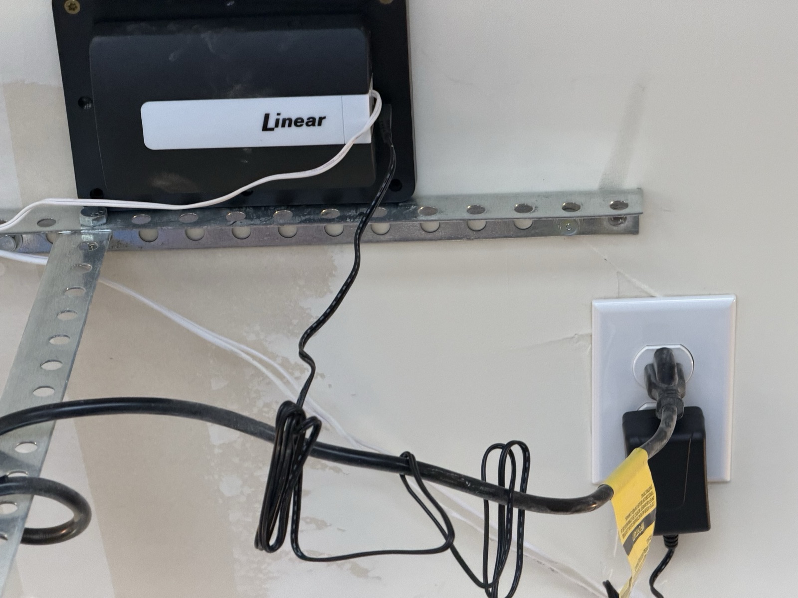 Linear-branded Z-Wave garage door controller module mounted on a wall, wired to a nearby power outlet with a low-voltage lead.