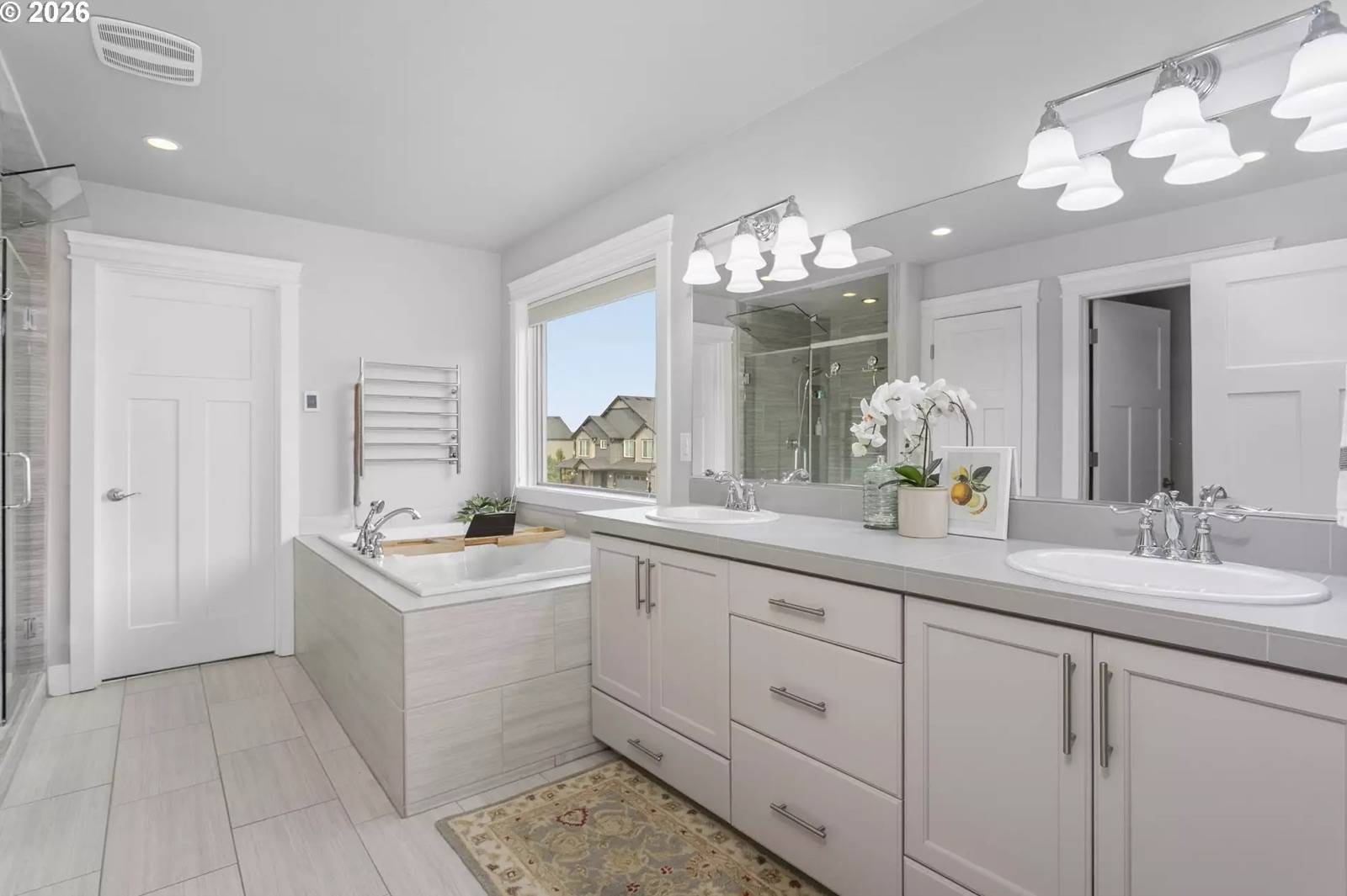 Wide view of the master bath: a tiled-surround soaking tub under a window, a chrome ladder-style heated towel rack on the wall beside it, a long double-sink vanity with a full-width mirror, large-format floor tile, and the glass shower enclosure at the left edge of the frame.