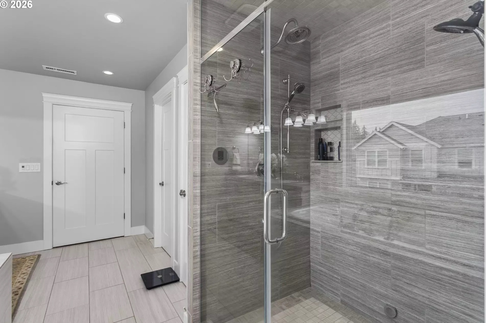 The master shower enclosure: a full-height glass front with a swing-out glass door and a long bar handle, stone-look tile to the ceiling, a rain head overhead, a wall head and a handheld sprayer on a slide bar, two valve trims, and a mosaic-backed niche.
