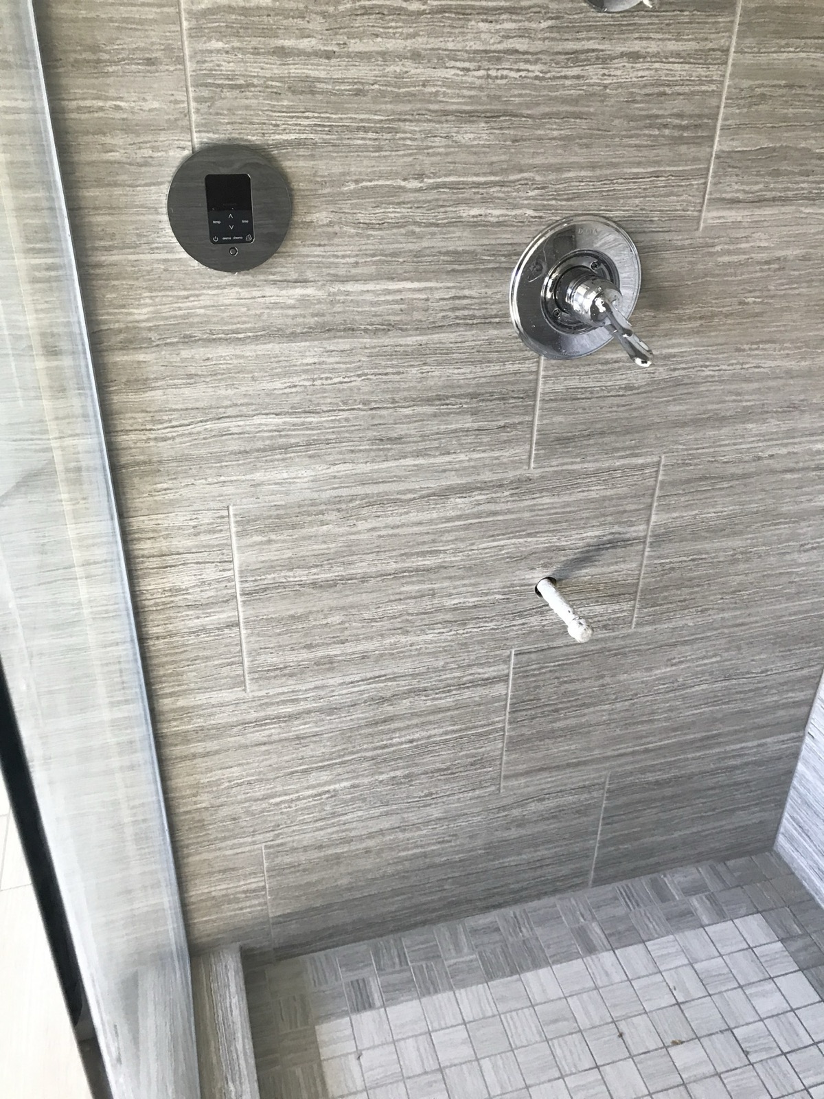The shower’s south wall: the round Mr. Steam iTempoPlus control with its display and temperature and time buttons, the shower valve trim to its right, and a low chrome outlet on the wall well below both, above the tiled floor.