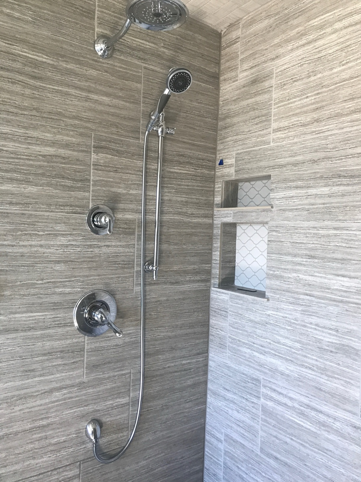 Tiled steam shower with a fixed rain shower head, a handheld sprayer on a slide bar, two separate valve trims, and two tiled mosaic-accent niches.