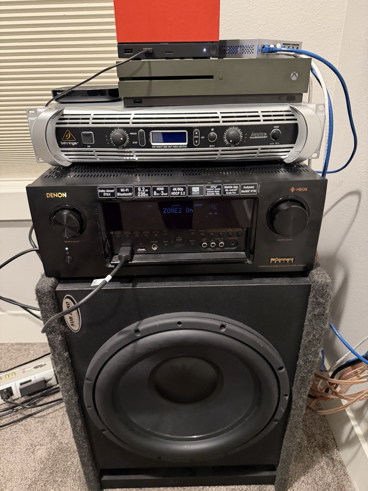 The media room electronics — a Roku, an Xbox, a small network switch, and an orange-faced Steam Machine gaming PC on top, the Behringer iNUKE NU3000DSP amp, the Denon AVR-X4300H receiver, and the Sub Zero Manufacturing subwoofer enclosure — sitting stacked on the carpet.