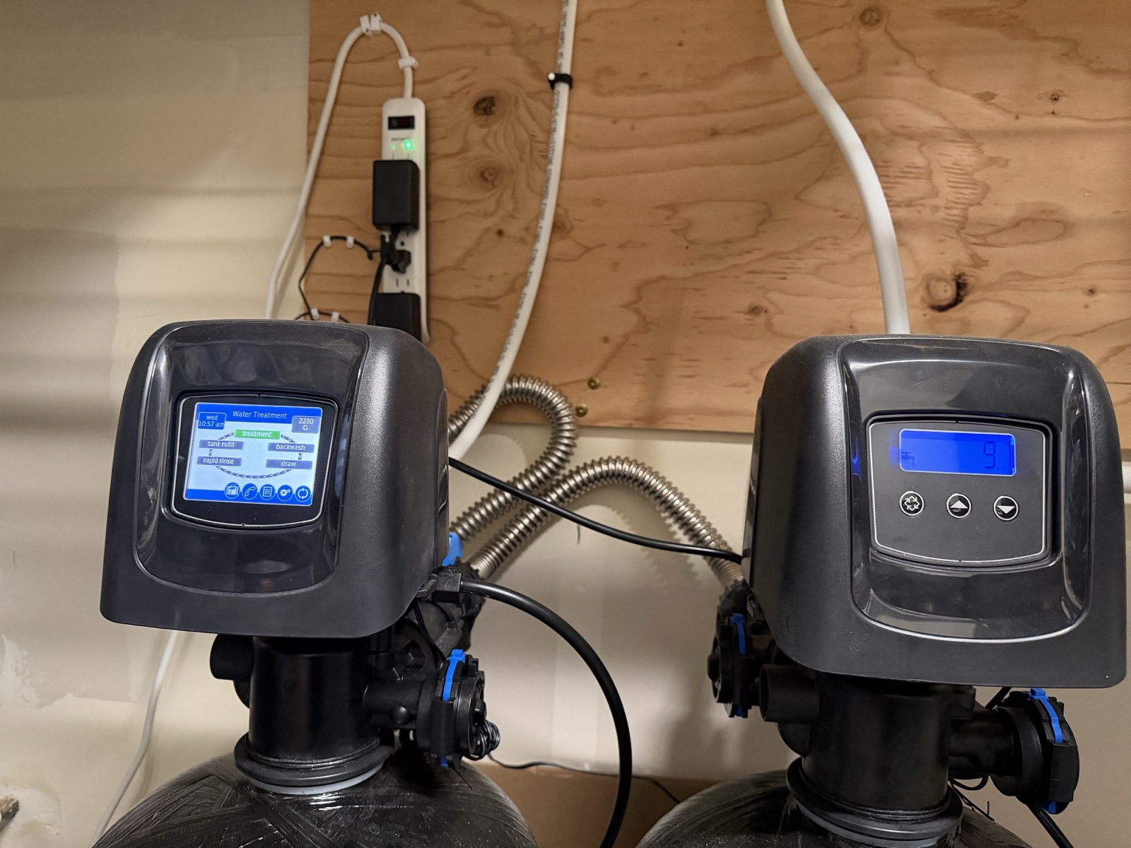 Two Clack WS1-series control valve heads on the water-treatment tanks — one with a color touchscreen, one with a blue-backlit three-button LCD — mounted below a plywood equipment backer board carrying a shared power strip.