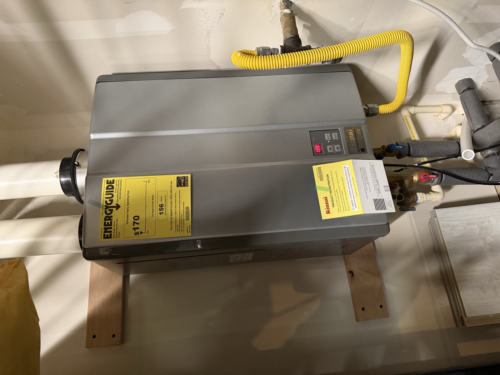 Rinnai RUC80i indoor condensing tankless gas water heater mounted on a wooden wall stand, with its yellow EnergyGuide label, yellow CSST gas line, and red/blue isolation valves.
