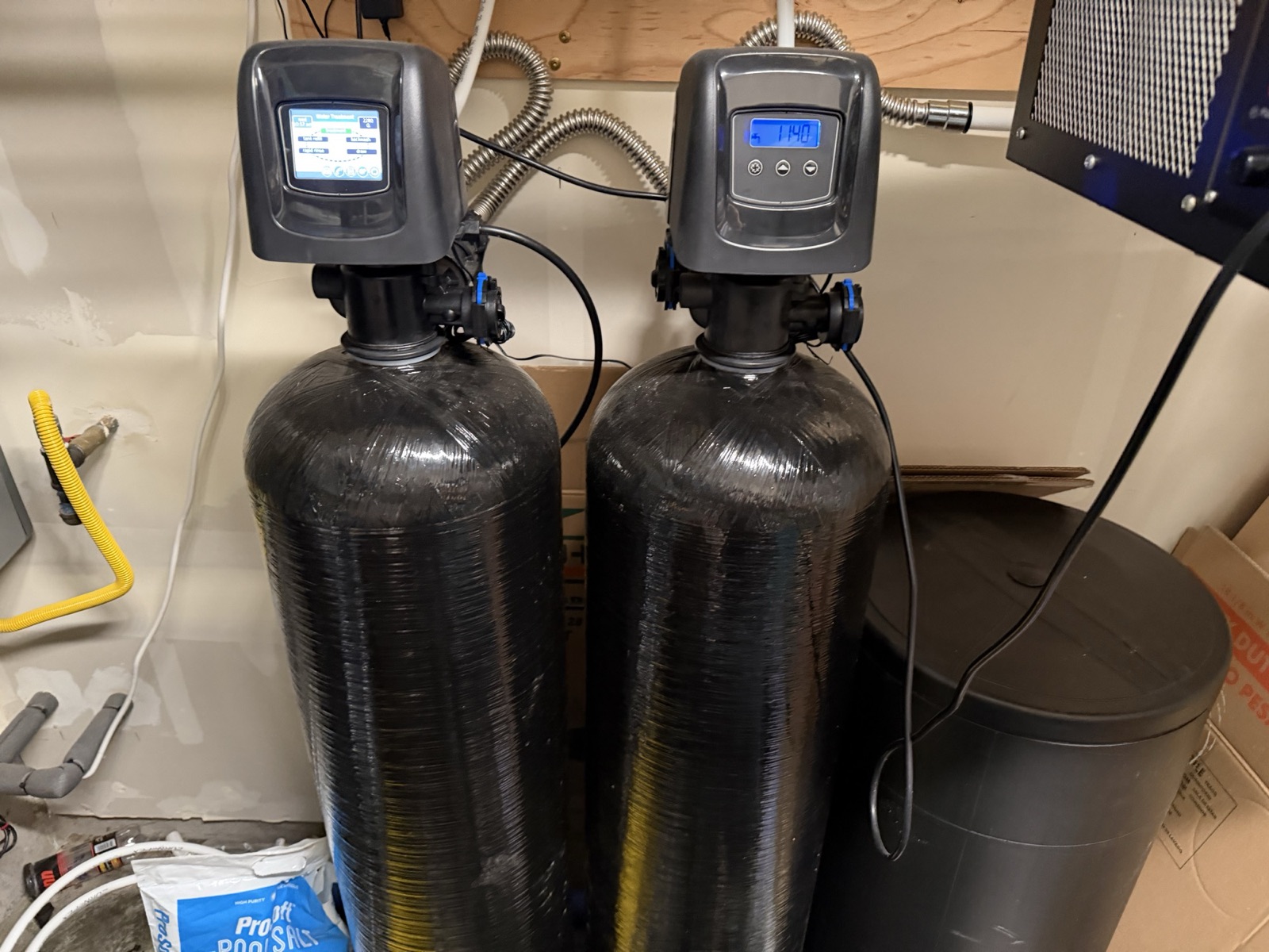 Wide view of the garage back bay: twin black water-treatment tanks with their control heads, the black brine tank and stacked salt bags at right, a yellow CSST gas stub in the wall, and the underside of a blue shop air-filtration unit hanging at the upper right, cut off by the frame.