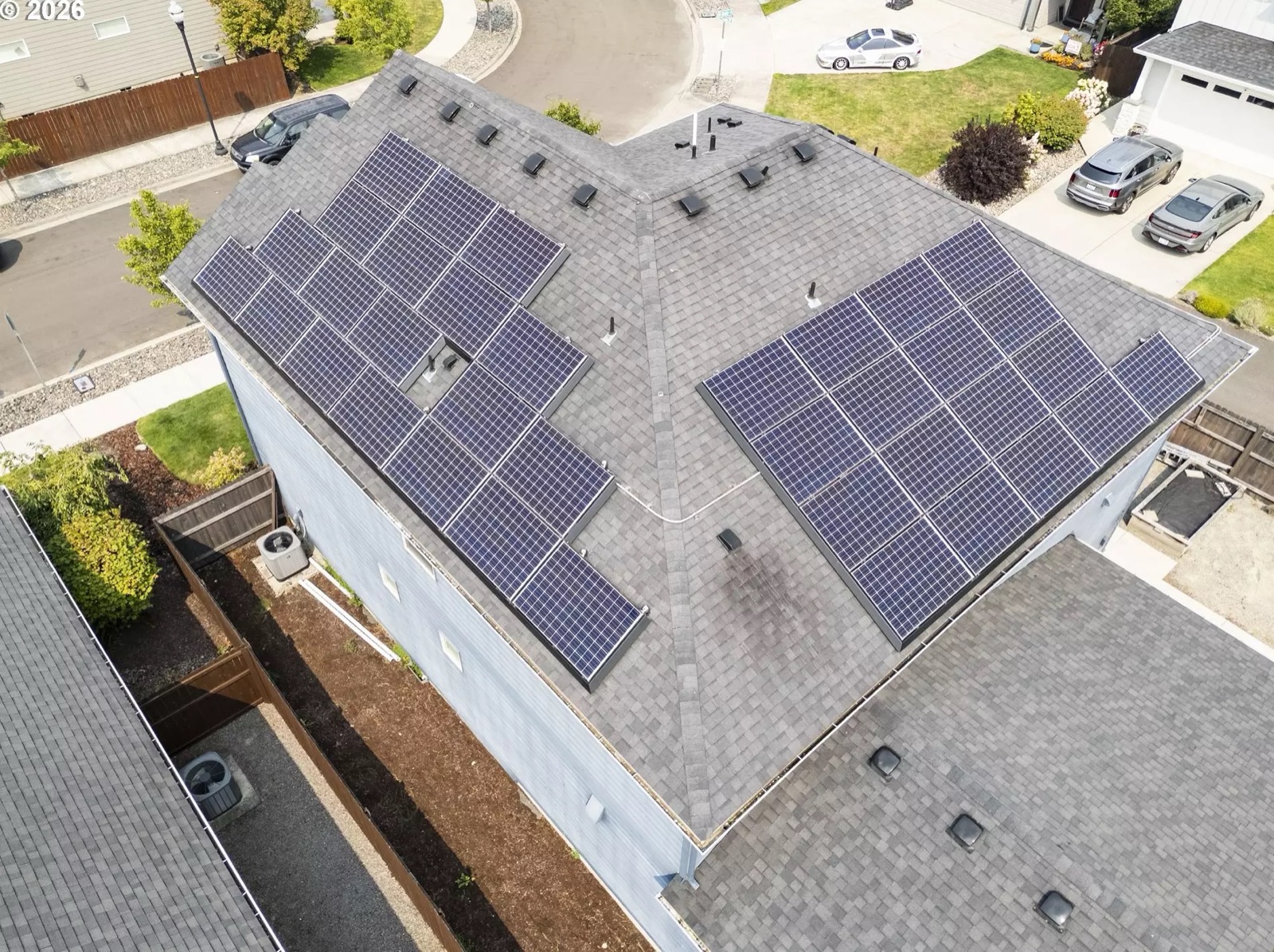 Close aerial view straight down onto the roof: two large blocks of blue photovoltaic modules laid out on two adjoining roof planes either side of the hip, with roof vents between them and an outdoor air-conditioning condenser on its pad against the side wall below.