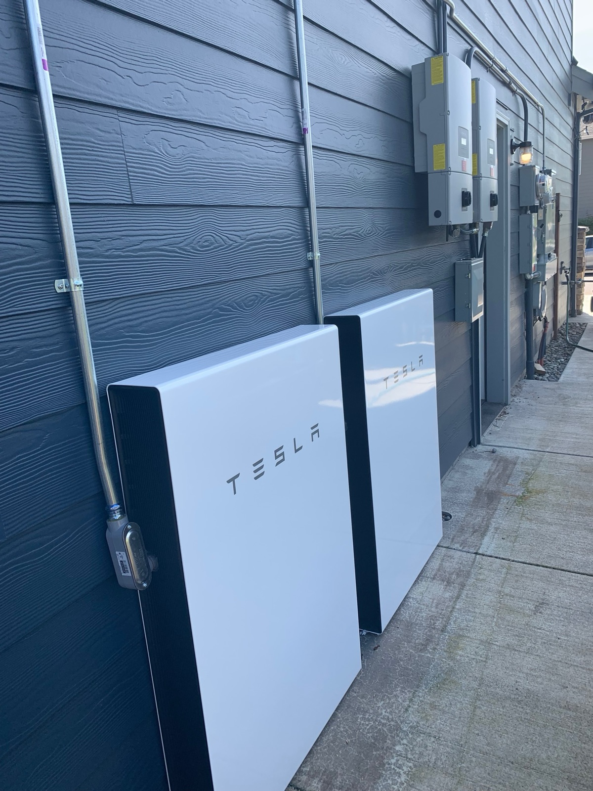 Two white Tesla Powerwall batteries standing against the side of the house, with conduit and grey inverter enclosures on the wall above them.