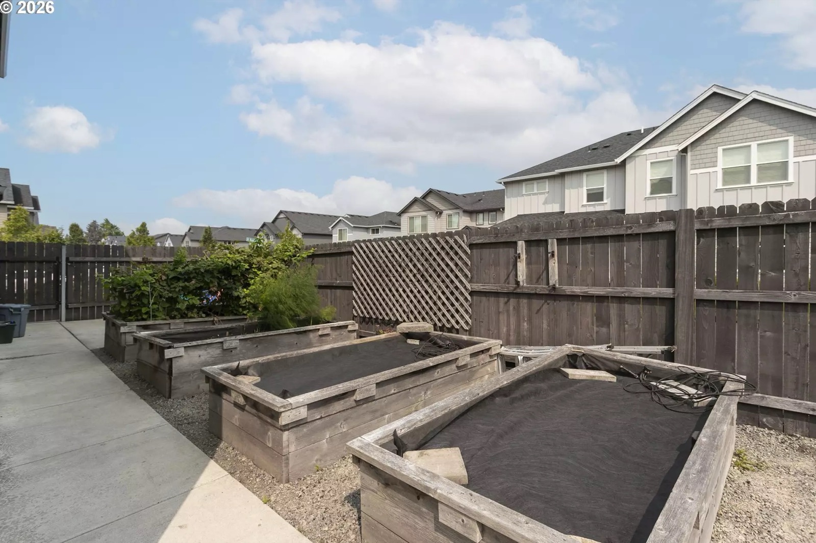 Three timber raised garden beds on a gravel bed beside a concrete path, each covered with dark fabric, against a wood fence with a lattice panel and a gate.